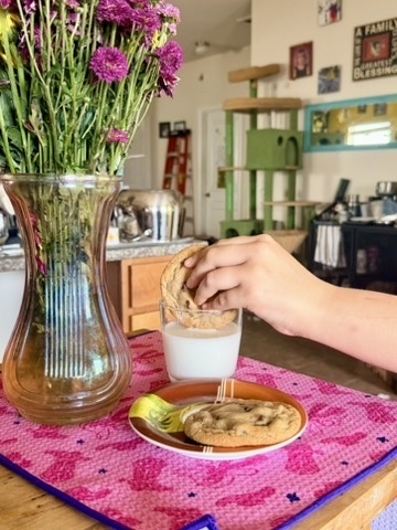 A chocolate chip cookie being dipped into milk beside fresh flowers